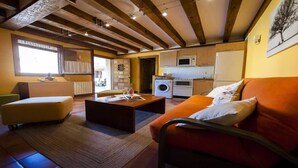 A cozy living room with a sofa, ottoman, and coffee table. There is a kitchen area with appliances and a window. - Villa Don Juan II (Segovia)