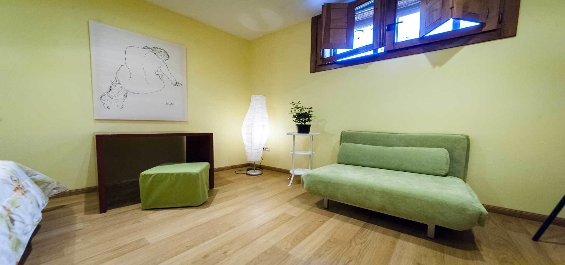 A room with a green sofa, a small green ottoman, a lamp, a painting, and a window with blinds.