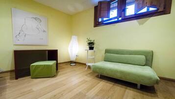A room with a green sofa, a small green ottoman, a lamp, a painting, and a window with blinds.