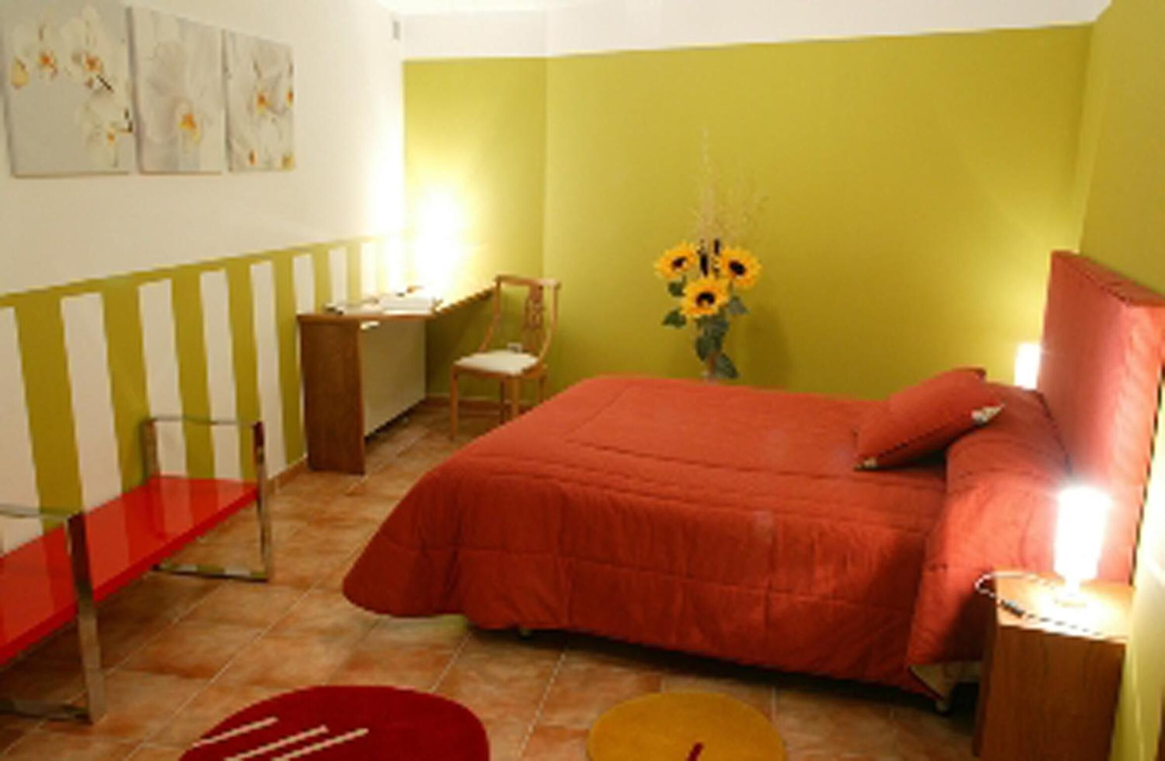 Un dormitorio con cama, escritorio, silla y un florero con girasoles.