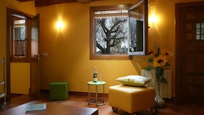 A cozy room with a wooden bench, a small table with a bottle and glass, a green stool, and a window with a view of trees. - Villa Don Juan II (Segovia)
