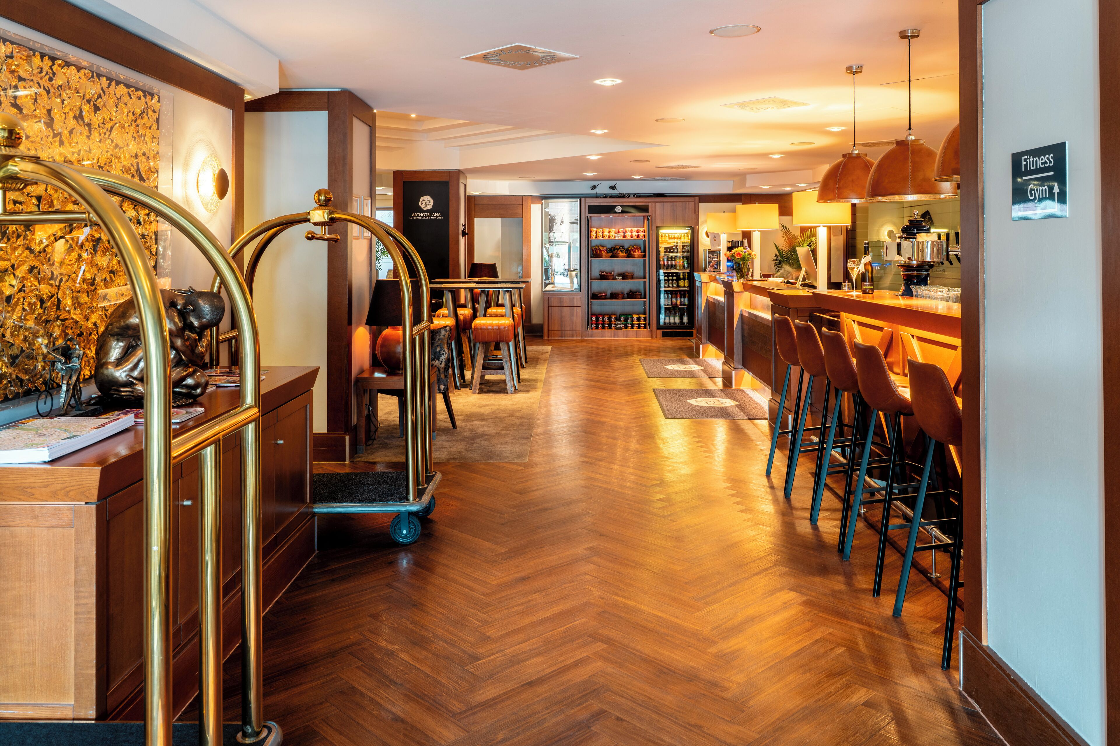 Un hall d’hôtel avec un parquet en bois disposé en chevrons, un comptoir d’accueil, un coin bar avec des tabourets et un panneau indiquant l’emplacement du centre de remise en forme.