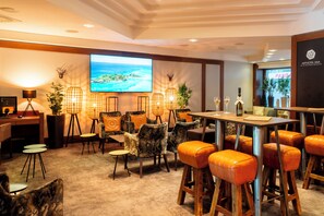 A hotel lobby with a large screen displaying a scenic view, multiple seating areas with cushioned chairs, a bar area with stools, and a reception desk. - Arthotel ANA im Olympiapark (Munich)