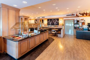 A modern kitchen with a central island, wooden cabinets, and a built-in stove. There is a seating area with a chair and a small table. - Arthotel ANA im Olympiapark (Munich)