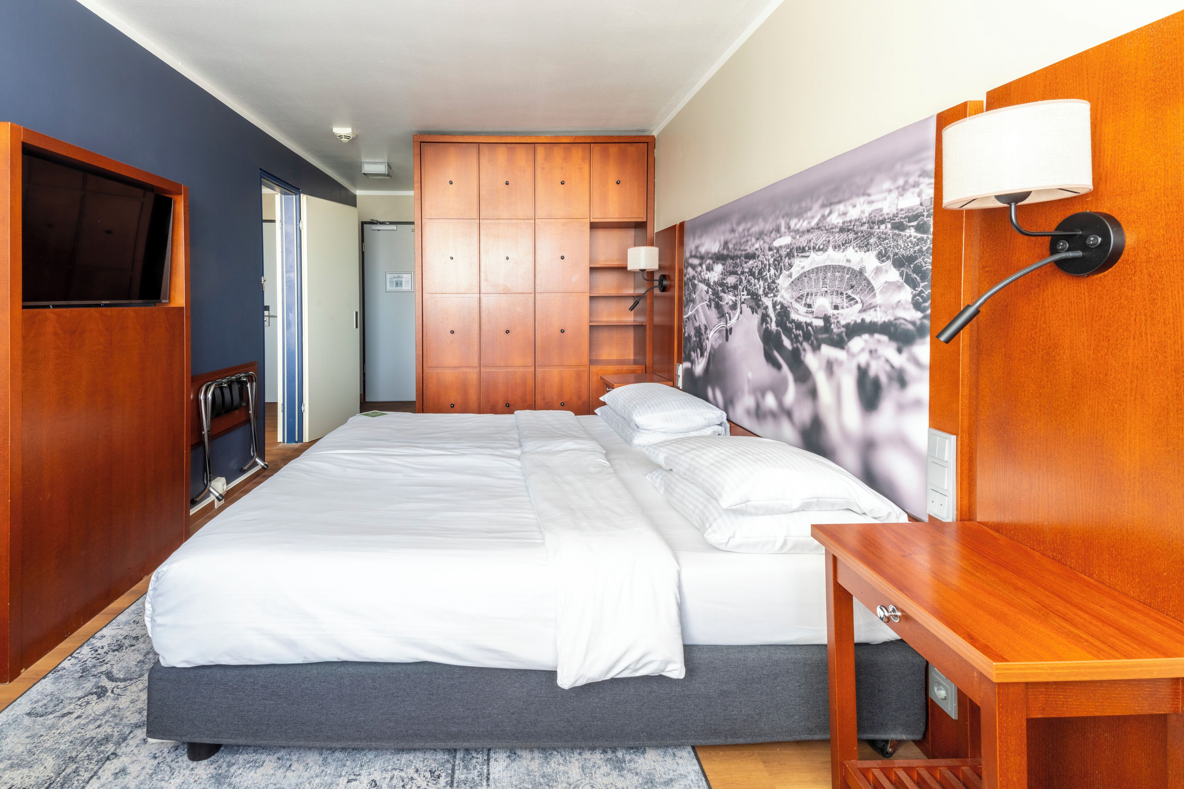Un dormitorio con una cama, un armario de madera, un televisor, una lámpara de pared y un cuadro de gran formato con una vista urbana.