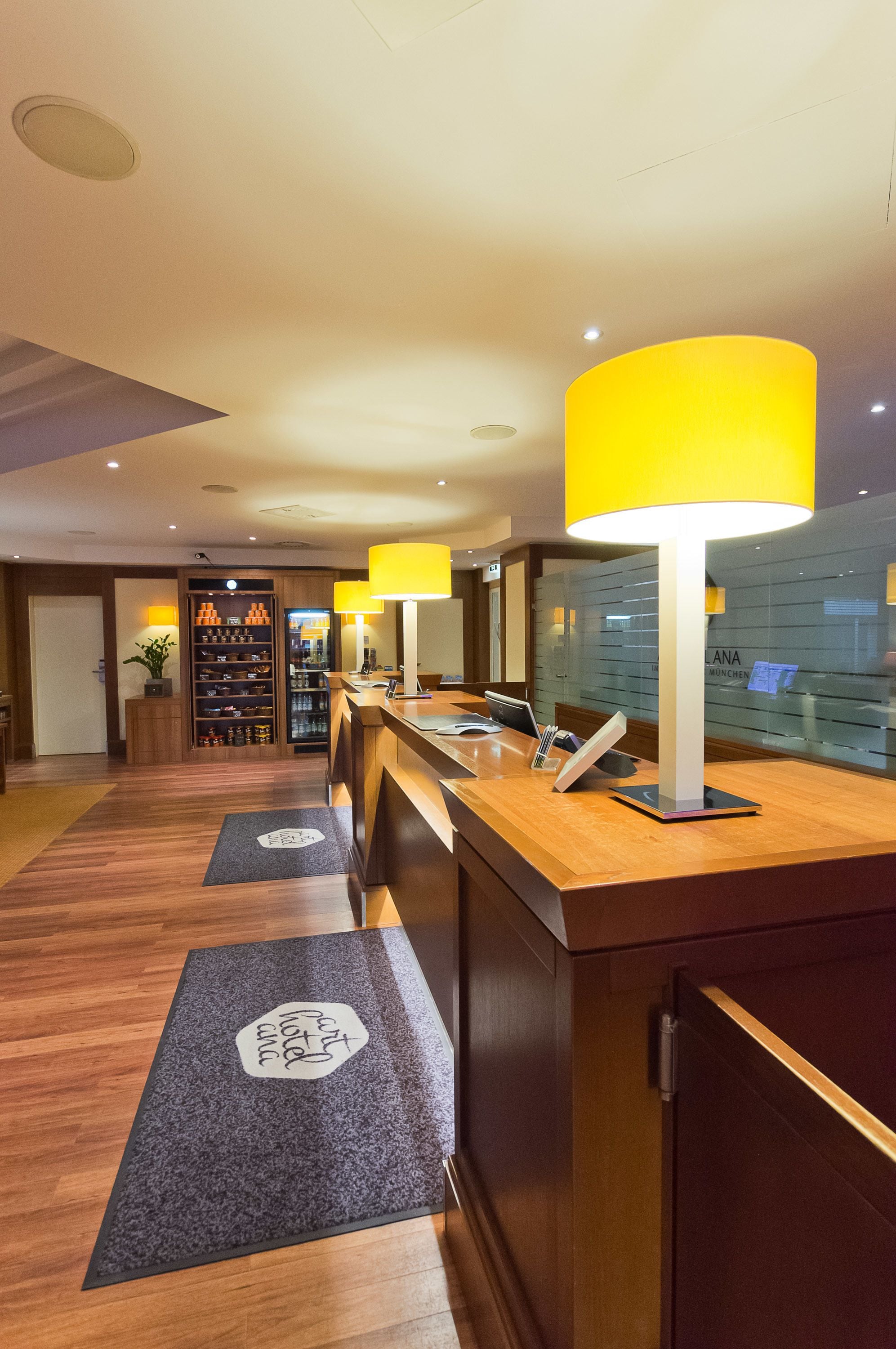 Et hotellets reception område med en træreception, en stor gul lampe, et skoshowroom og en glasvæg med refleksioner.
