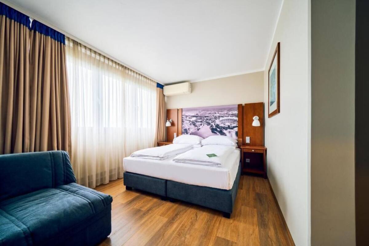 Ein Hotelzimmer mit einem Bett, einem Sofa, einem großen Fenster mit Vorhängen, einem Gemälde an der Wand und einem Nachttisch mit einer Lampe.