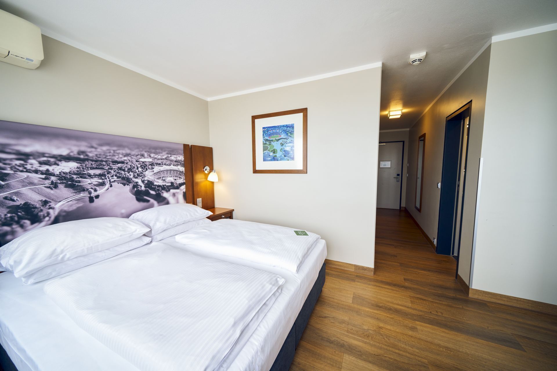 A hotel room with a large bed, a nightstand with a lamp, a framed picture on the wall, and a hallway leading to another room.