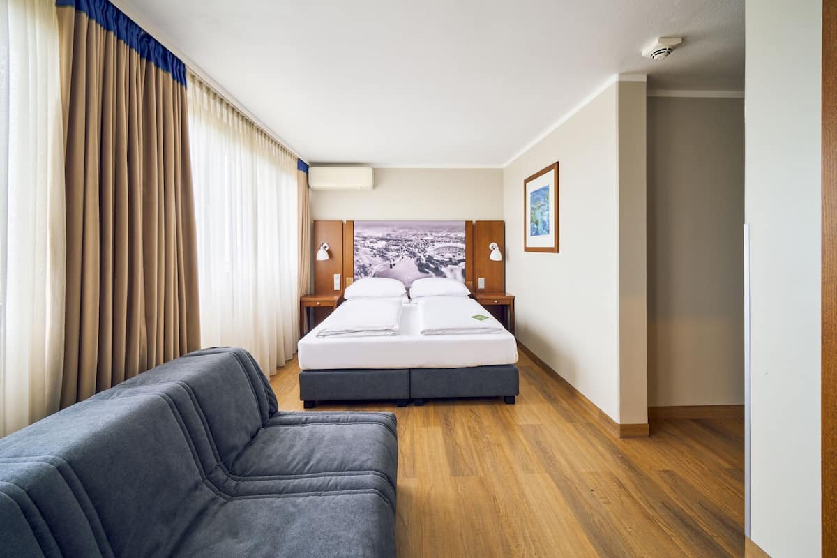 Ein Hotelzimmer mit einem Bett, einem Schreibtisch, einem Sessel und einem großen Fenster mit Vorhängen.