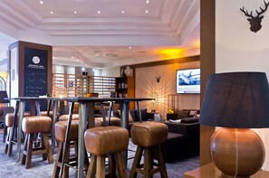 A modern hotel lobby with leather bar stools, a reception desk, a large screen displaying a news channel, and a wall-mounted deer head decoration. - Arthotel ANA im Olympiapark (Munich)