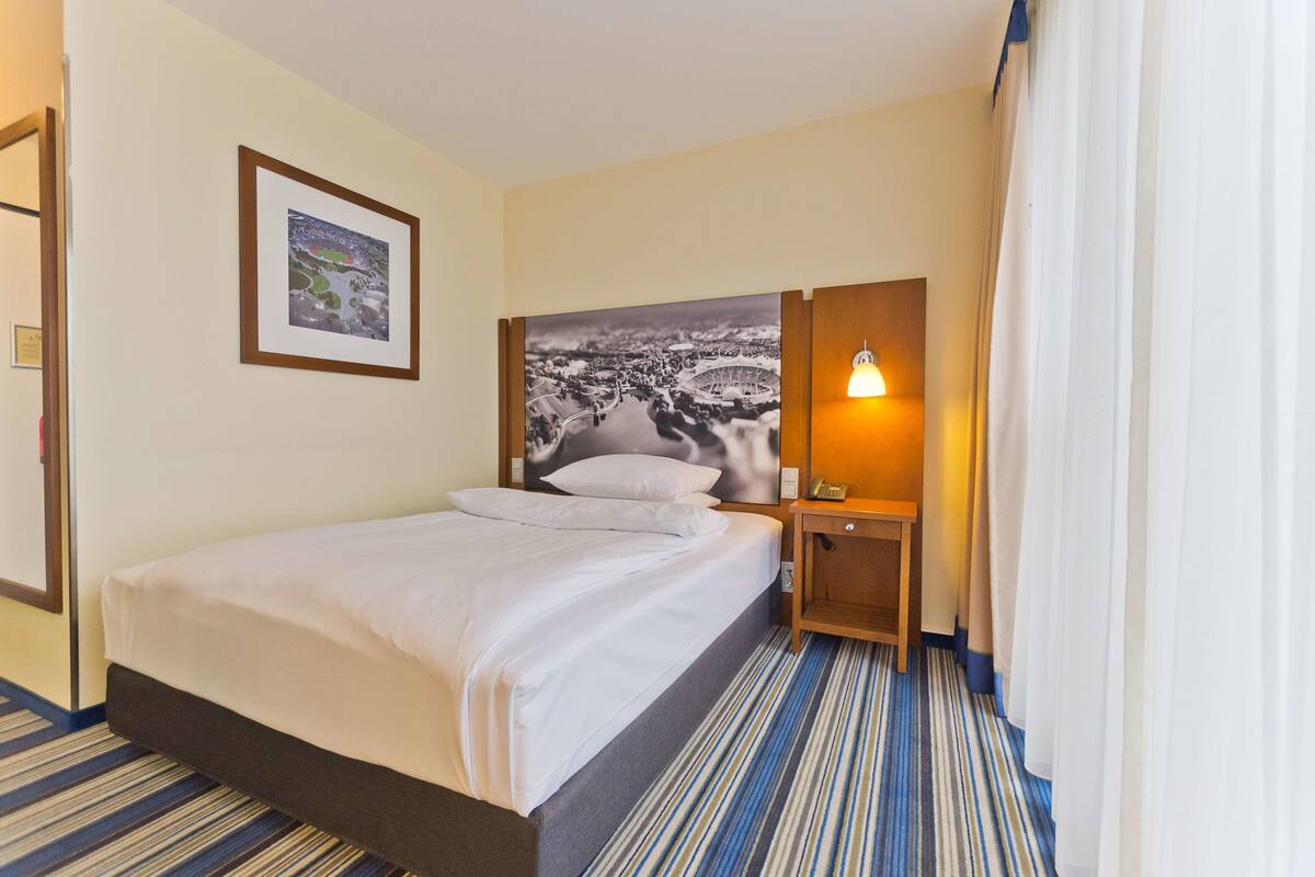 Ein Hotelzimmer mit Bett, einem gerahmten Bild an der Wand, einem Nachttisch mit Lampe und einem Streifenmuster-Teppich.