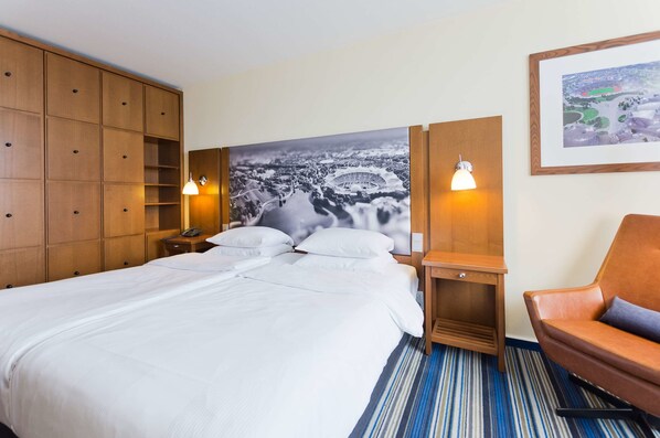 A hotel room with a large bed, wooden wardrobe, two bedside tables with lamps, a chair, and a large framed picture on the wall. - Arthotel ANA im Olympiapark (Munich)