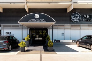 The entrance to ARTHOTEL ANA with a covered entrance, two potted plants, and a parking area with cars. - Arthotel ANA im Olympiapark (Munich)