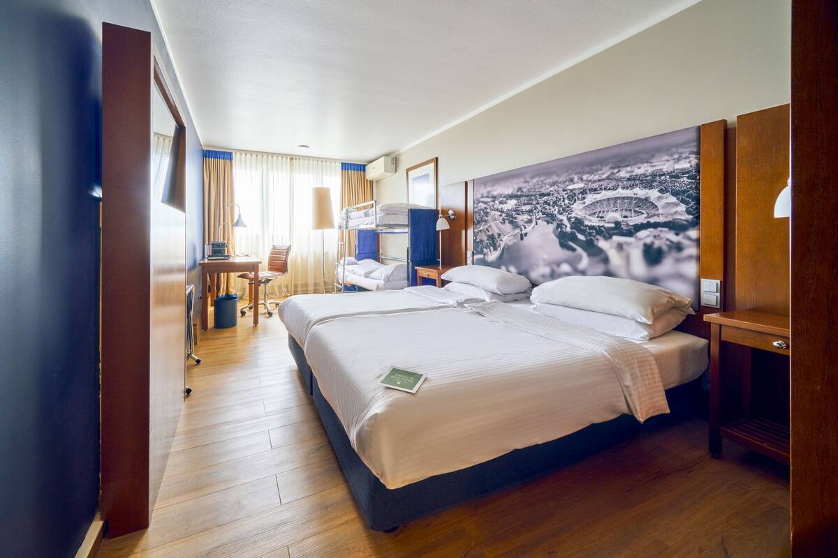 Ein Hotelzimmer mit einem großen Bett, einem Schreibtisch, einem Sessel und Blick auf ein Stadion durch das Fenster.
