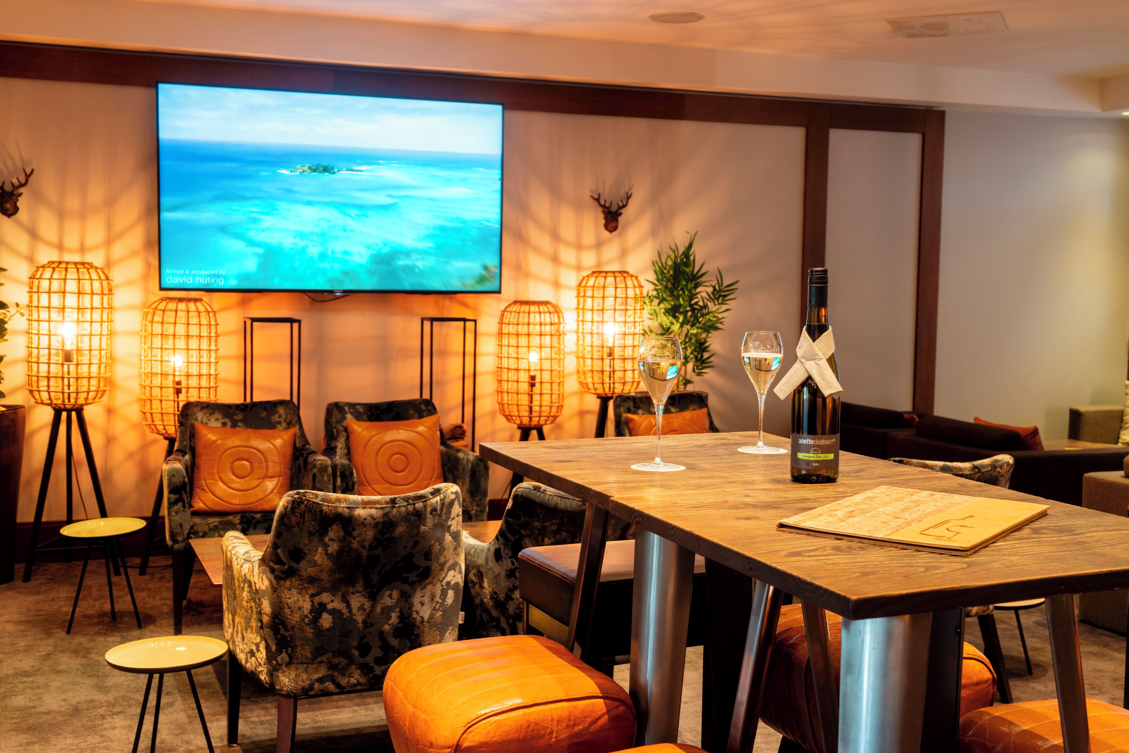 A hotel room with a large TV displaying a scenic ocean view, a wooden dining table with wine glasses, and a cozy seating area with patterned chairs and orange cushions.