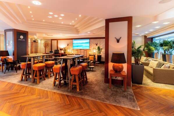 A spacious lobby with a wooden floor, a bar area with stools, a sofa, and a large TV screen. - Arthotel ANA im Olympiapark (Munich)