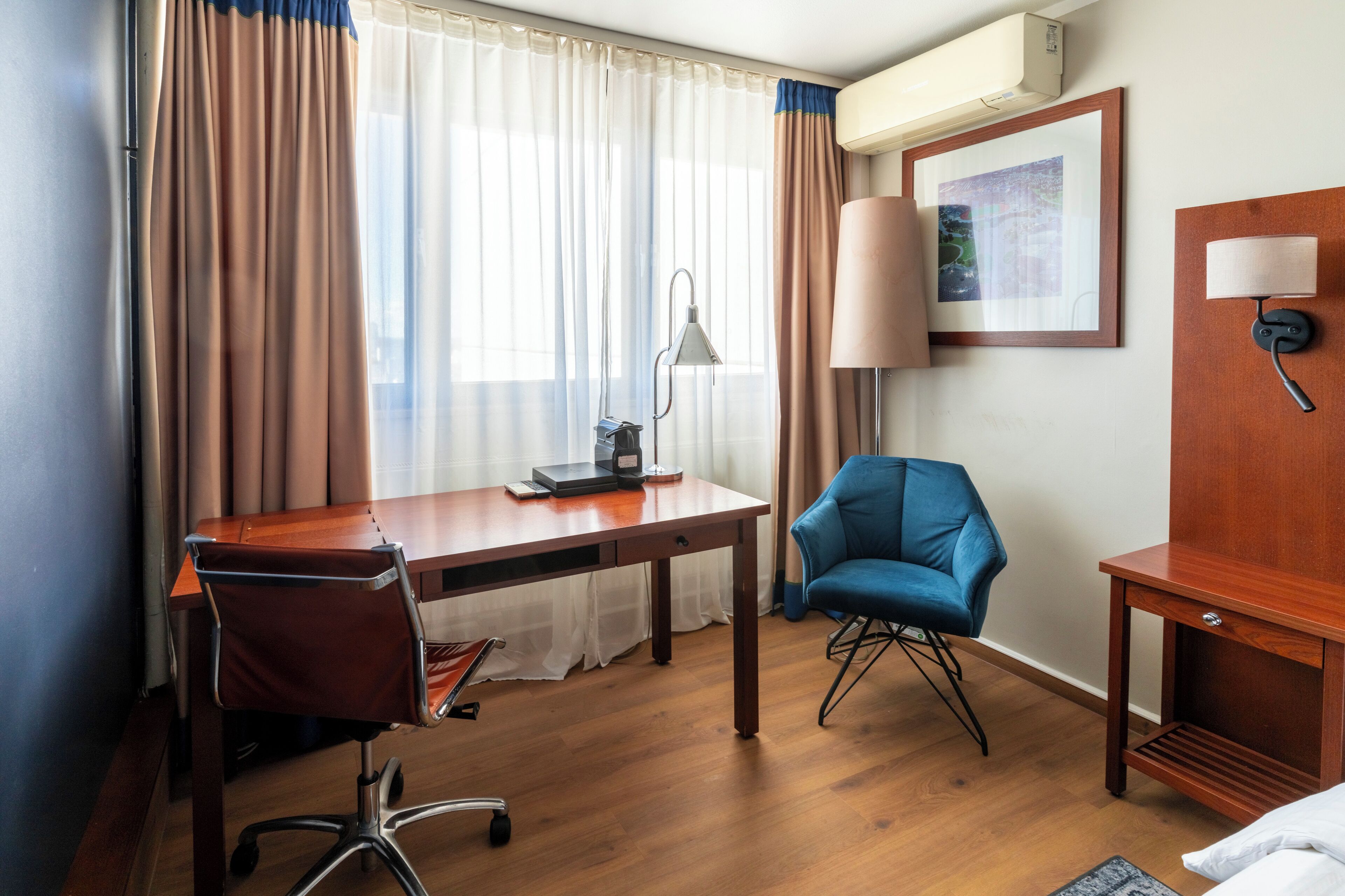 Una habitación de hotel con un escritorio de madera, una silla azul, una lámpara y una cama.
