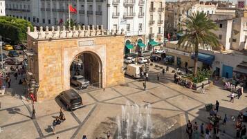 A public square with a large archway, fountains, and people walking around.
