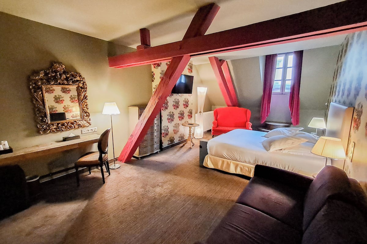A hotel room with a bed, a desk, a chair, a red armchair, and a mirror.