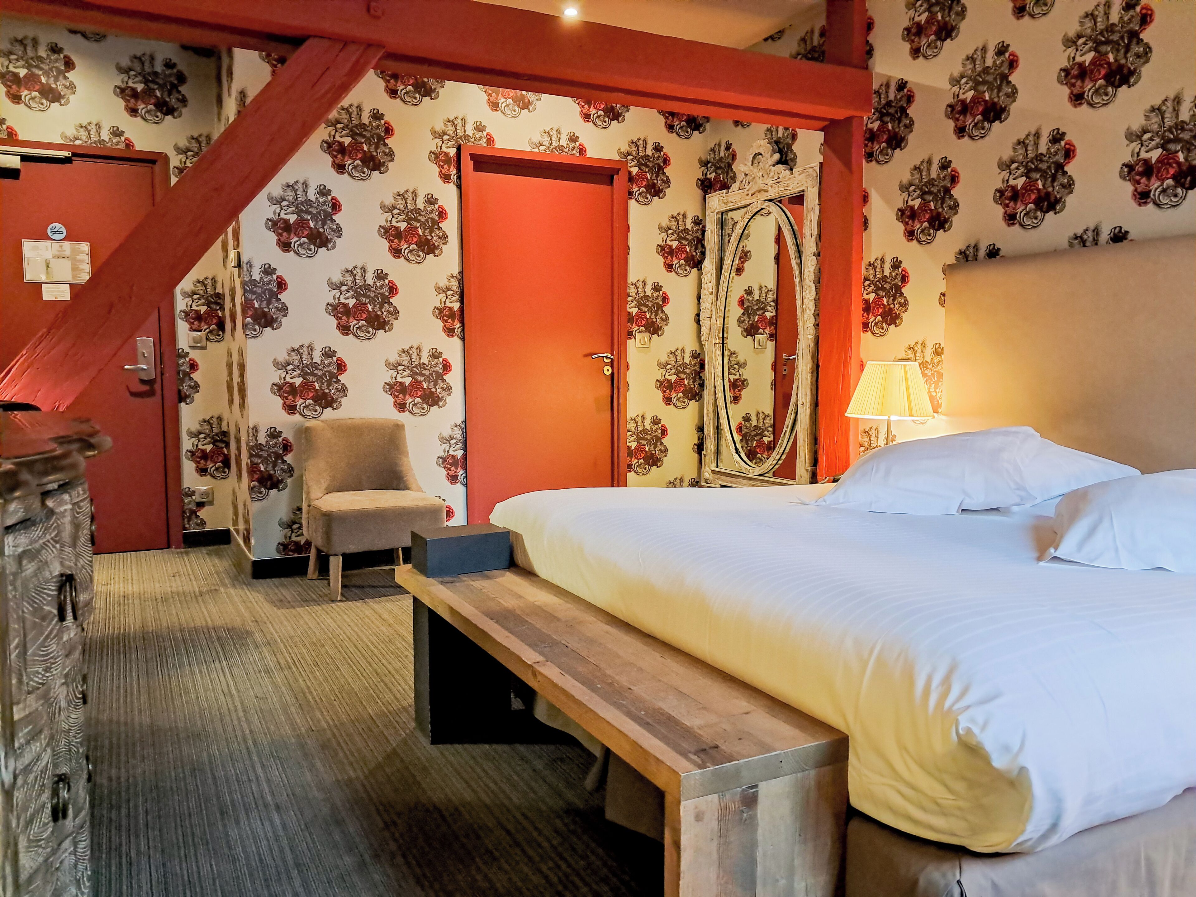 Une chambre d’hôtel avec un lit, une chaise, un banc, un miroir et une porte recouverte d’un papier peint à motifs floraux.