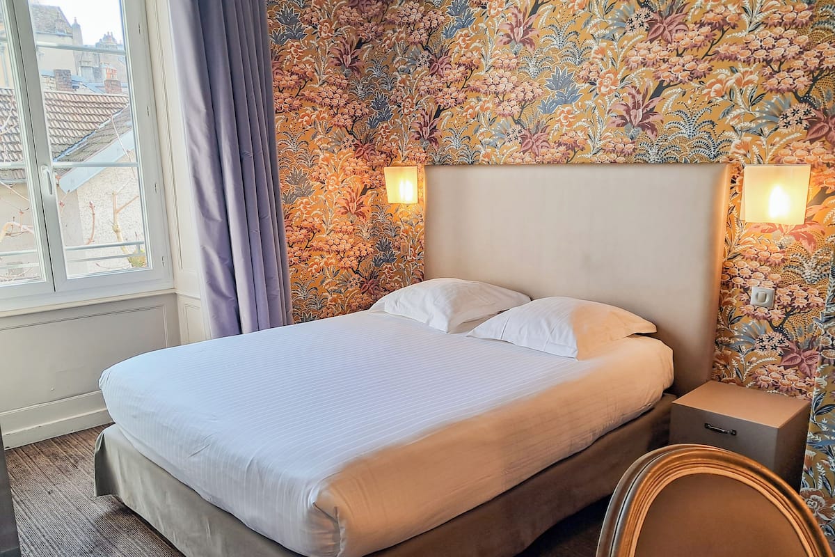 A neatly made bed with white linens, a floral-patterned wall, and a window with curtains.