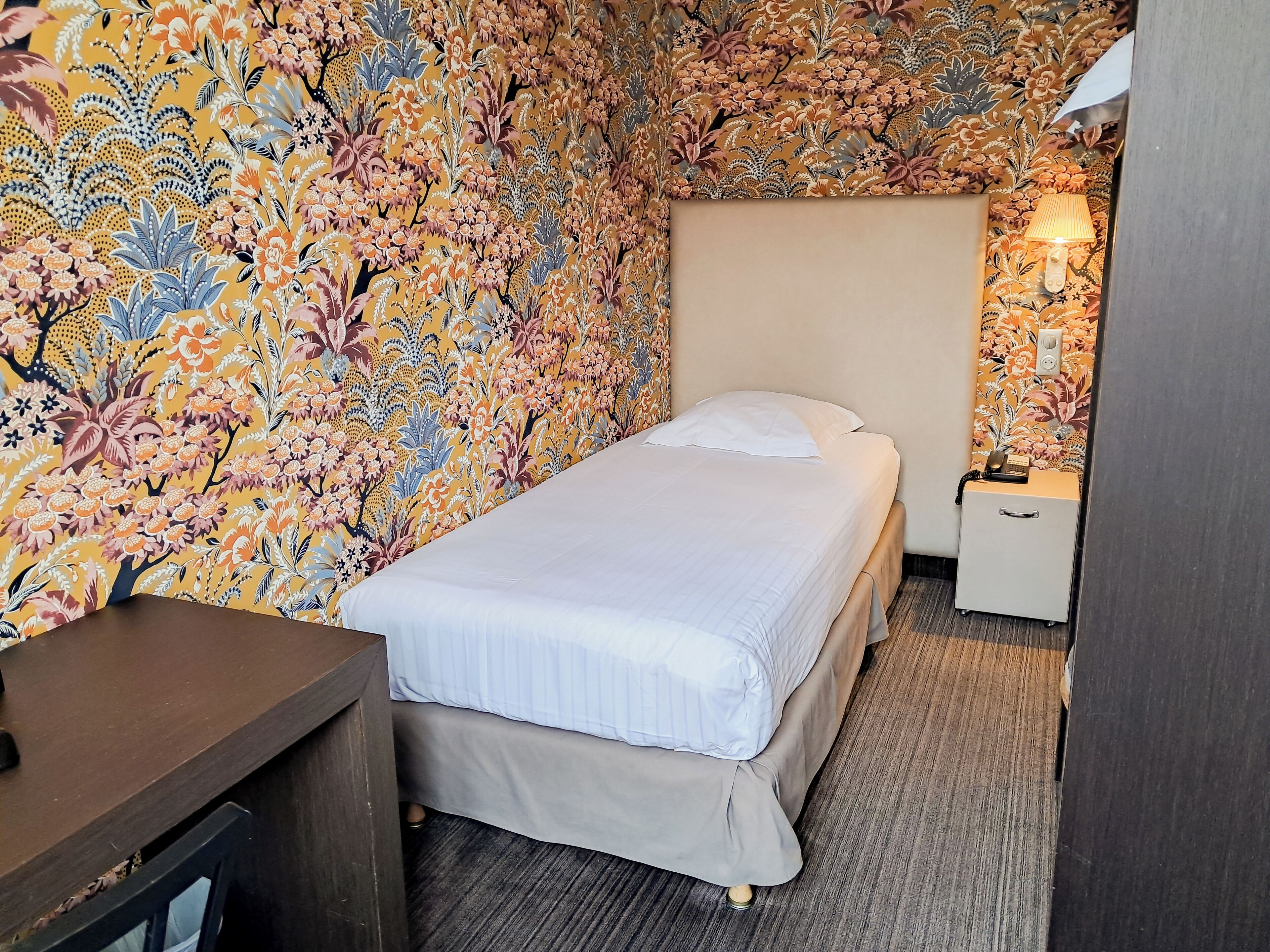 A small, neatly arranged room with a bed, a bedside table, a lamp, and a telephone.