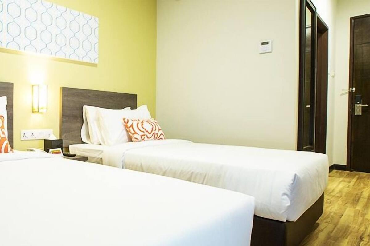 Two single beds with white linens and orange patterned pillows, a nightstand with a lamp, and a door with a lock.