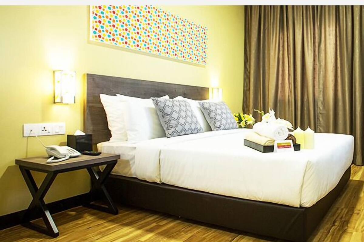 A hotel room with a bed, bedside table, lamp, TV, and a desk with a kettle and bottles.
