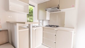 A compact kitchenette with a gas stove, a sink, and a window providing a view of greenery outside. - Camping Les Chèvrefeuilles (Royan)