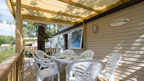 A covered outdoor patio with a wooden deck, white plastic chairs, and a table set for dining. - Camping Les Chèvrefeuilles (Royan)
