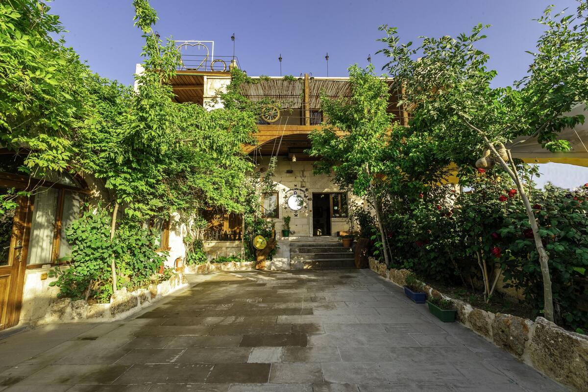 A courtyard with stone flooring, a wooden staircase leading to a building entrance, and lush greenery surrounding the area.