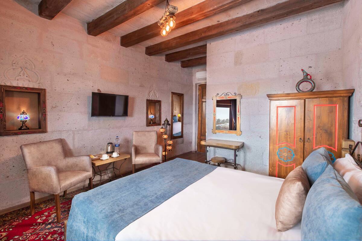 A bedroom with a bed, two chairs, a wooden wardrobe, a desk with a lamp, and a TV on the wall.