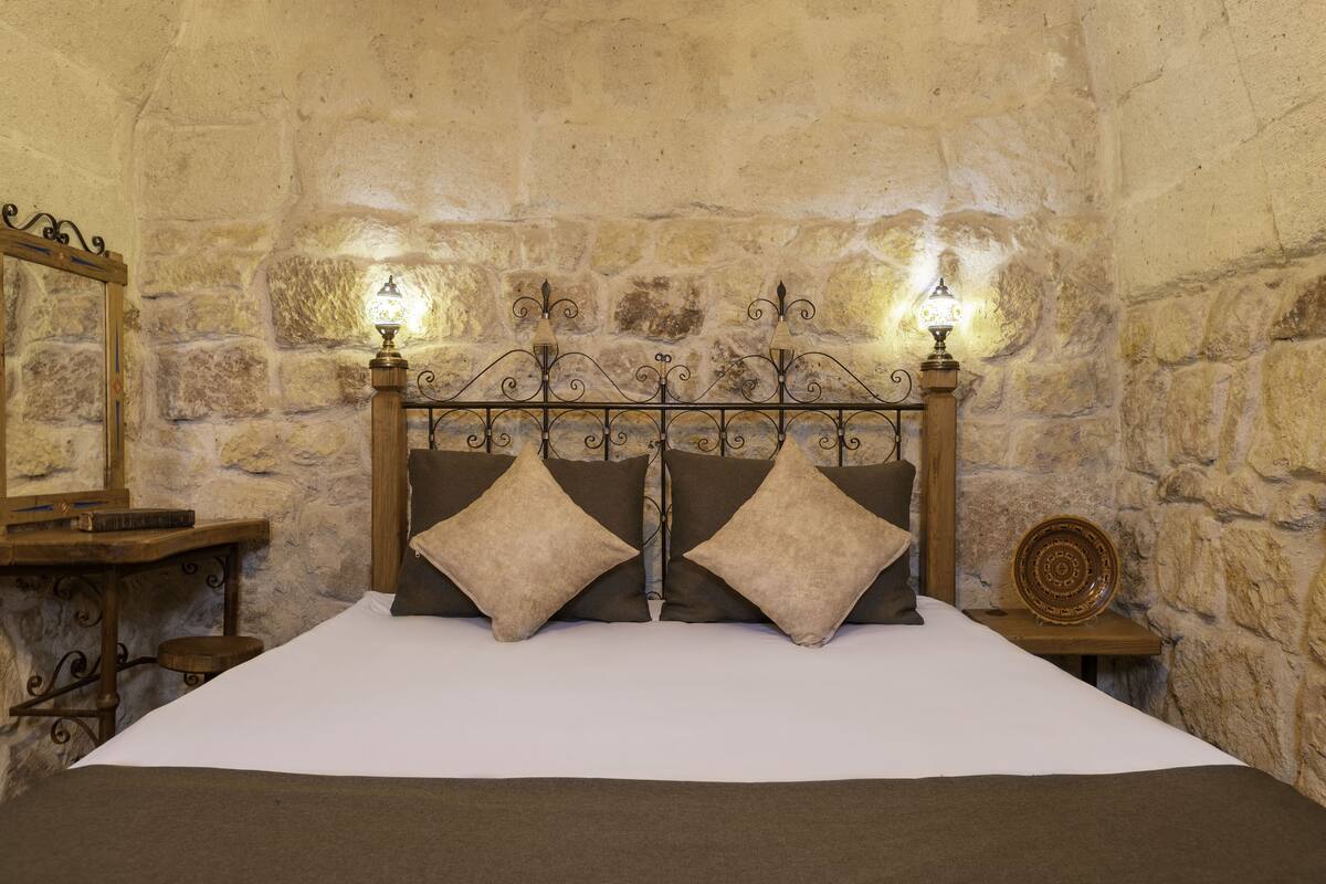 A bedroom with a stone wall, a wooden headboard, two bedside lamps, a mirror, and a small table with a decorative plate.