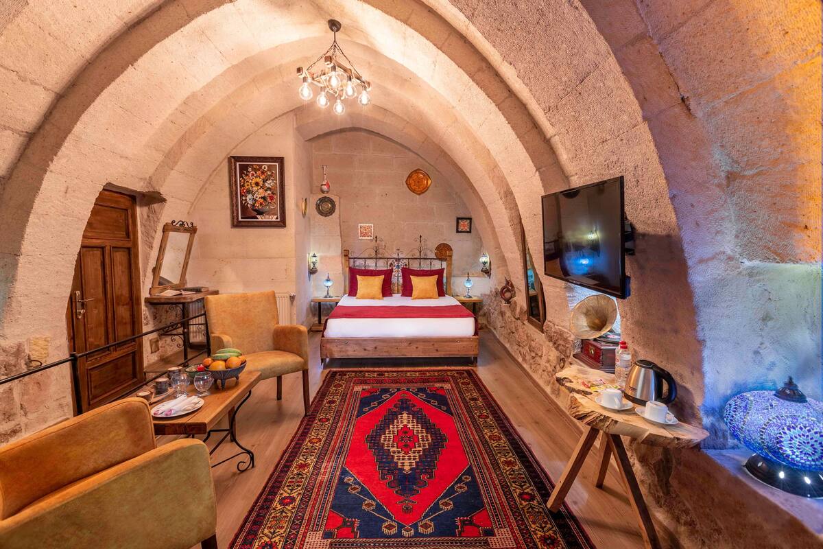 A cozy room with a wooden bed, a red and black patterned rug, a wooden table with a bowl of fruit, and a small table with a teapot and cups.