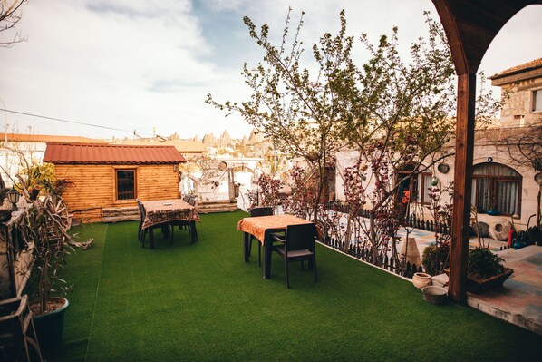 A courtyard with artificial grass, wooden tables, and chairs. There are potted plants and a small wooden building in the background. - Maron Stone House (Nevsehir)