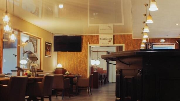 A restaurant with wooden flooring, multiple tables and chairs, hanging pendant lights, a flat-screen TV, and a bar area with stools. - Schlosspark-Hotel Hof von Oldenburg (Rastede)