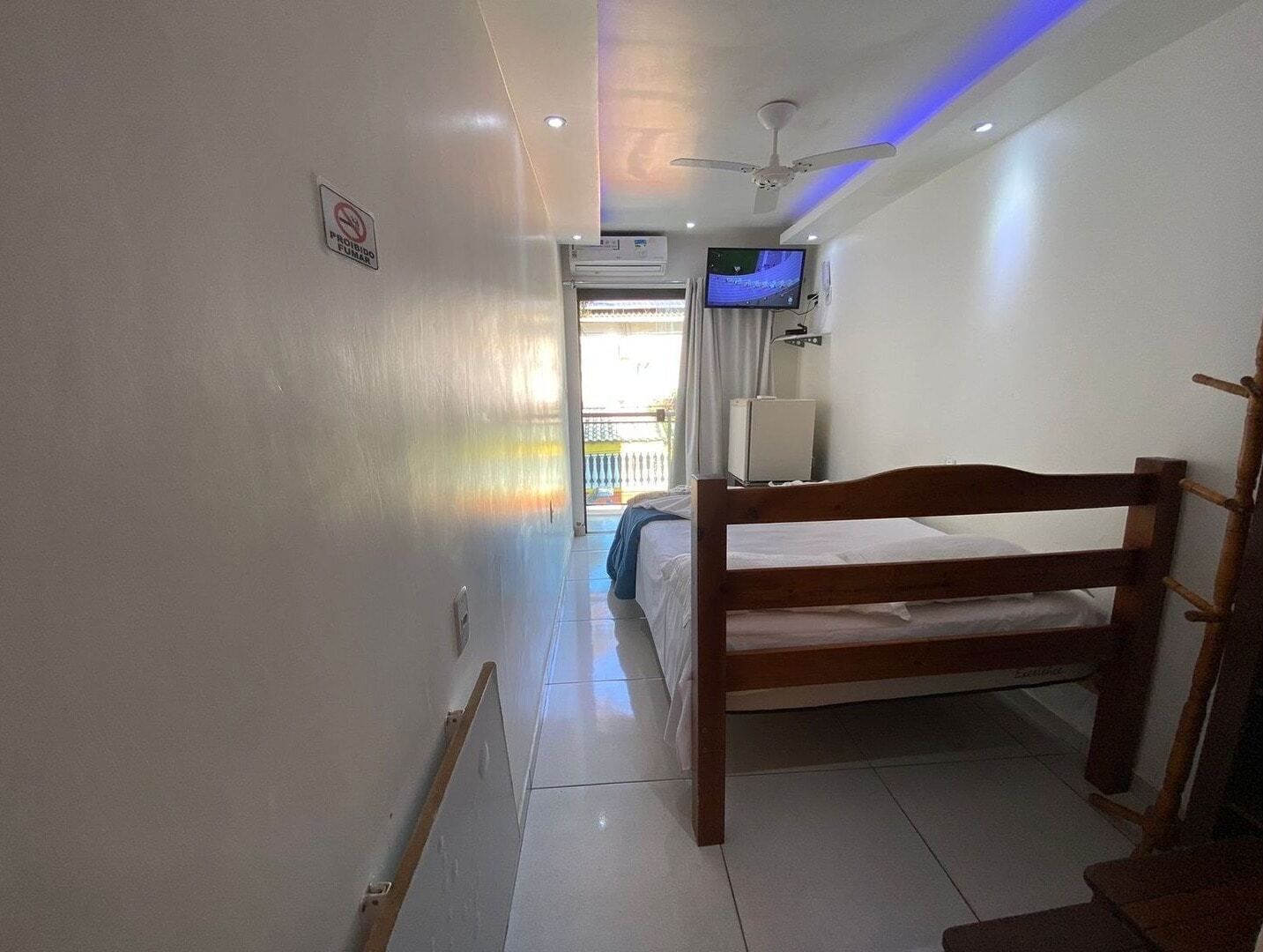 Habitació d'hotel amb un llit doble de fusta, un ventilador de sostre, una televisió penjada a la paret i una finestra amb cortines.