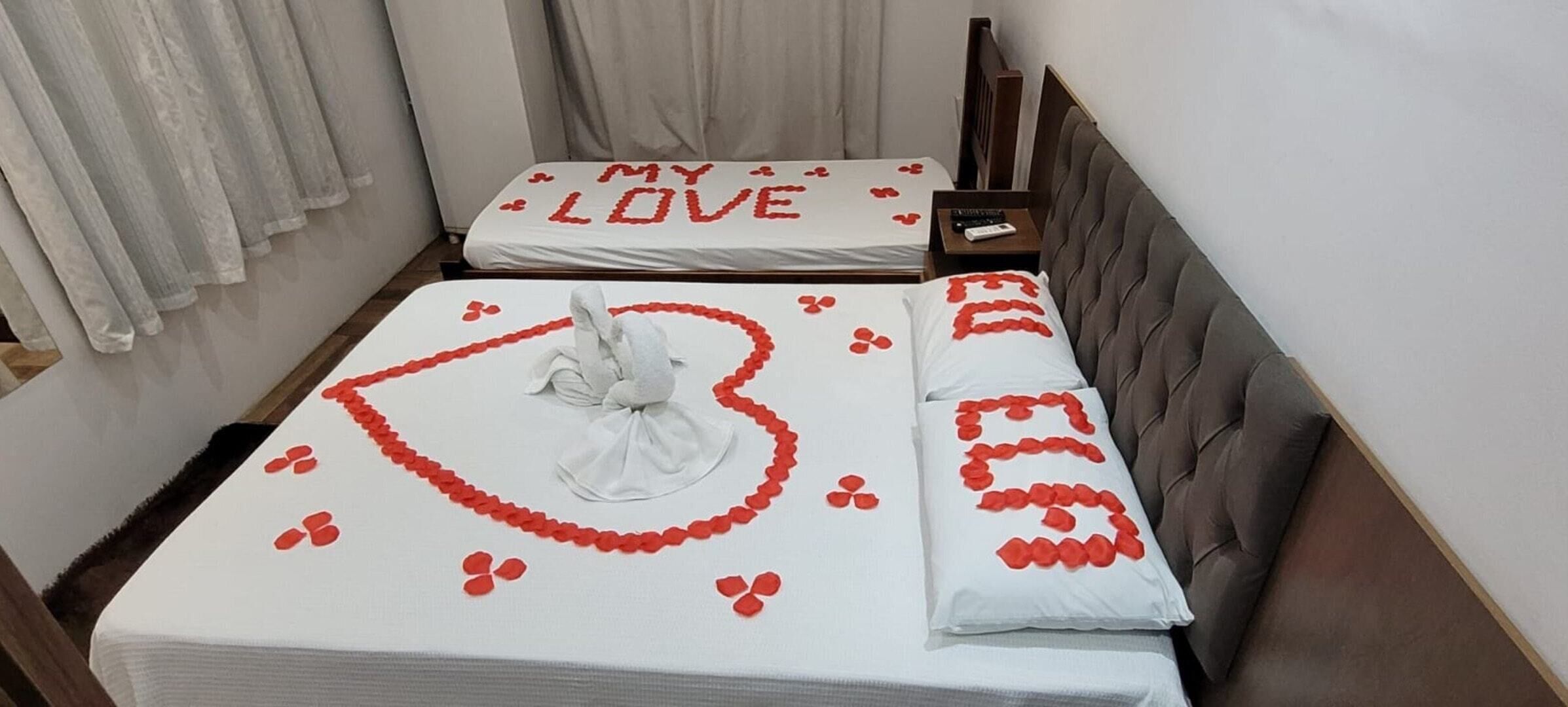 Habitación de hotel con dos camas, cada una adornada con un arreglo de pétalos de rosa roja en forma de corazón y una toalla blanca.