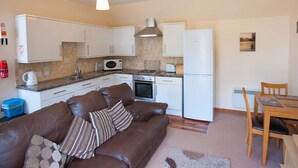 A compact kitchen with white cabinets, a microwave, and a stove. There's a brown leather sofa with pillows and a dining table with chairs.