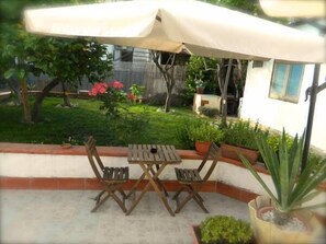 Une terrasse extérieure avec une table et des chaises sous un grand parasol, entourée de verdure et bordée d’un bâtiment blanc.