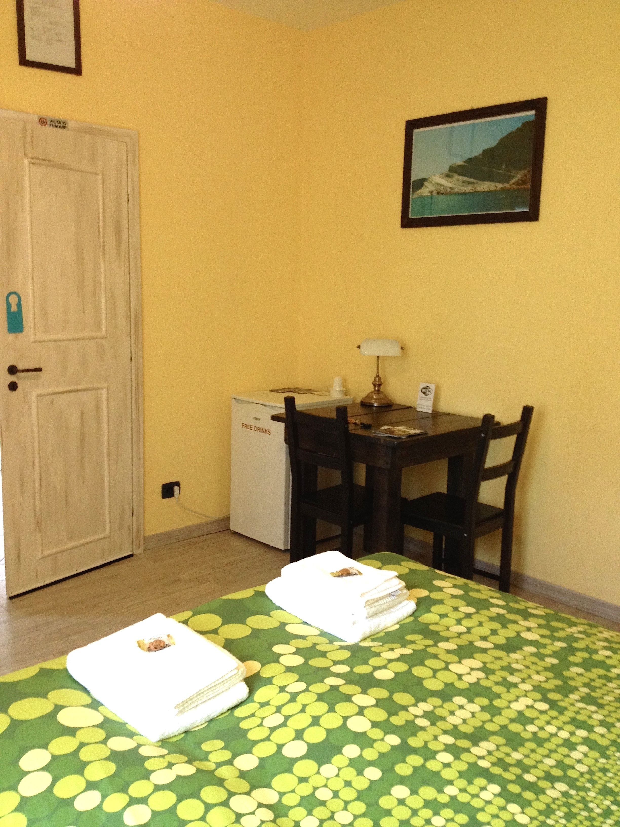 Una habitación con cama, escritorio, silla, un refrigerador pequeño y una puerta.