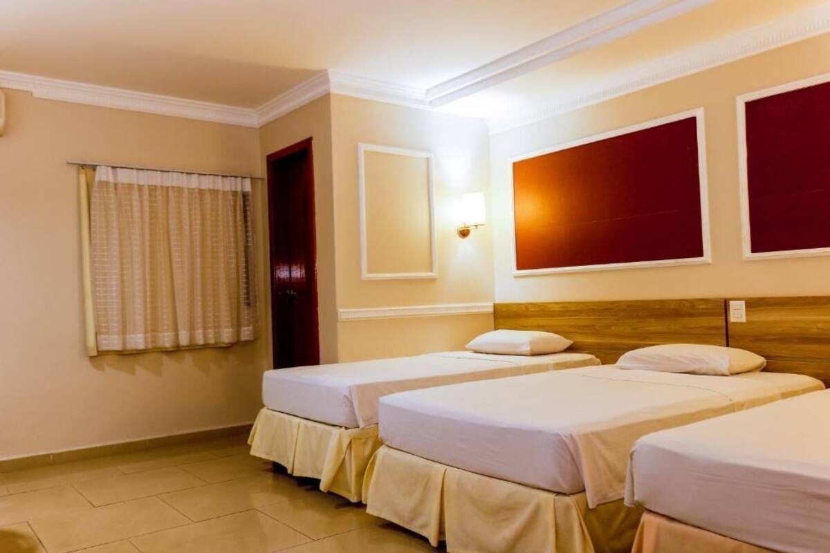 A hotel room with two single beds, a wooden headboard, a window with curtains, a wall-mounted light, and a door.