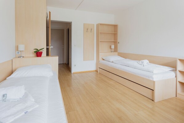 A modern hotel room with a wooden bed, a smaller bed, a nightstand with a lamp, a red plant, and a wooden wardrobe.