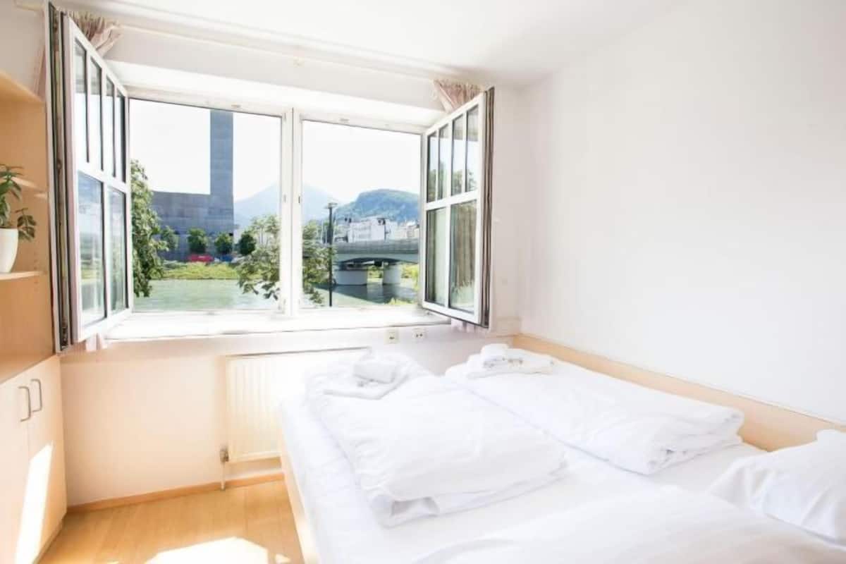 A hotel room with a large window, a bed with white linens, a radiator, and a potted plant.