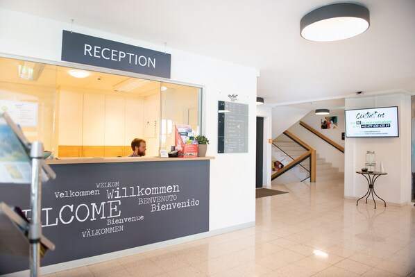 A reception area with a sign reading "RECEPTION" and a welcome sign with multiple languages. There is a staircase in the background and a table with a vase and a plant. - myNext - Riverside Hotel Salzburg (Salzburg)