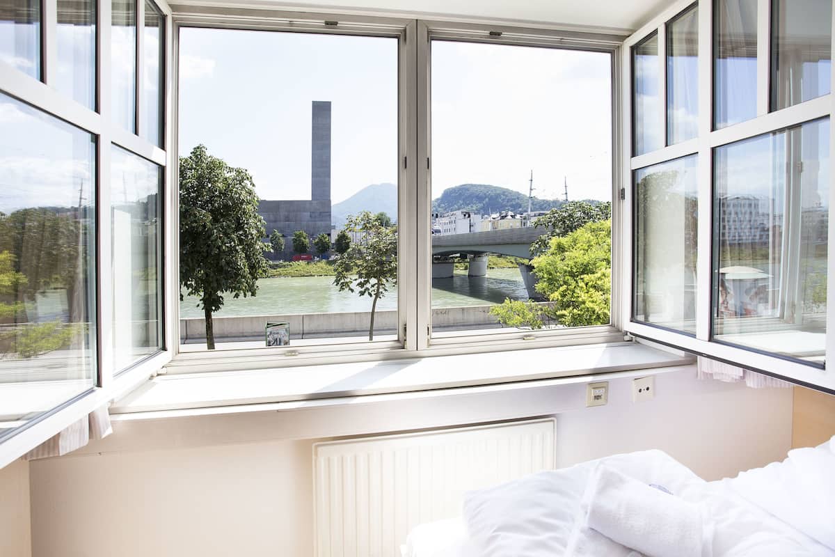 A bedroom with a large window overlooking a river, a bridge, and a mountain.