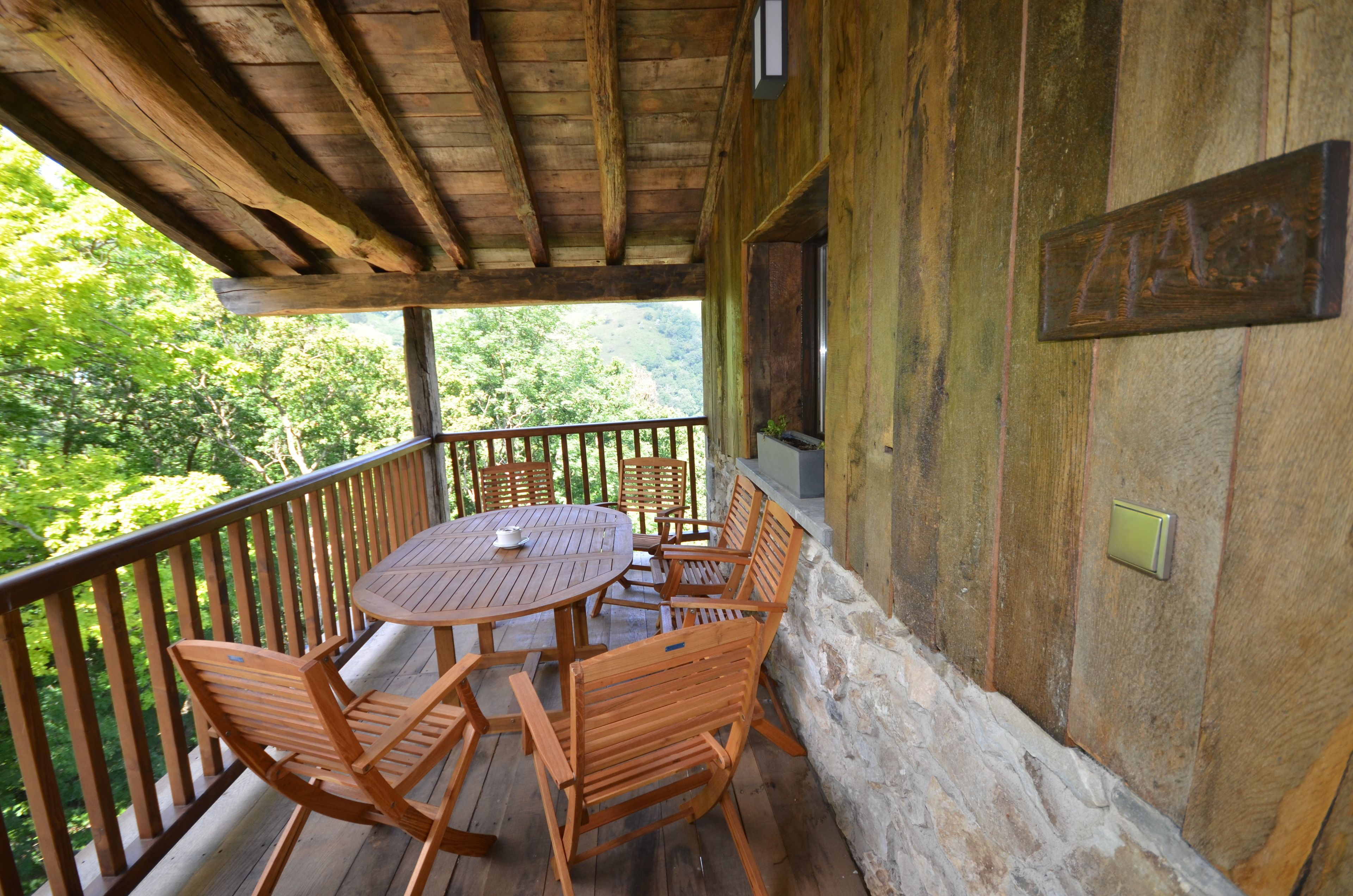 Une terrasse en bois avec une table ronde et des chaises, entourée d’un mur de pierre et d’une balustrade en bois, donnant sur une zone boisée.