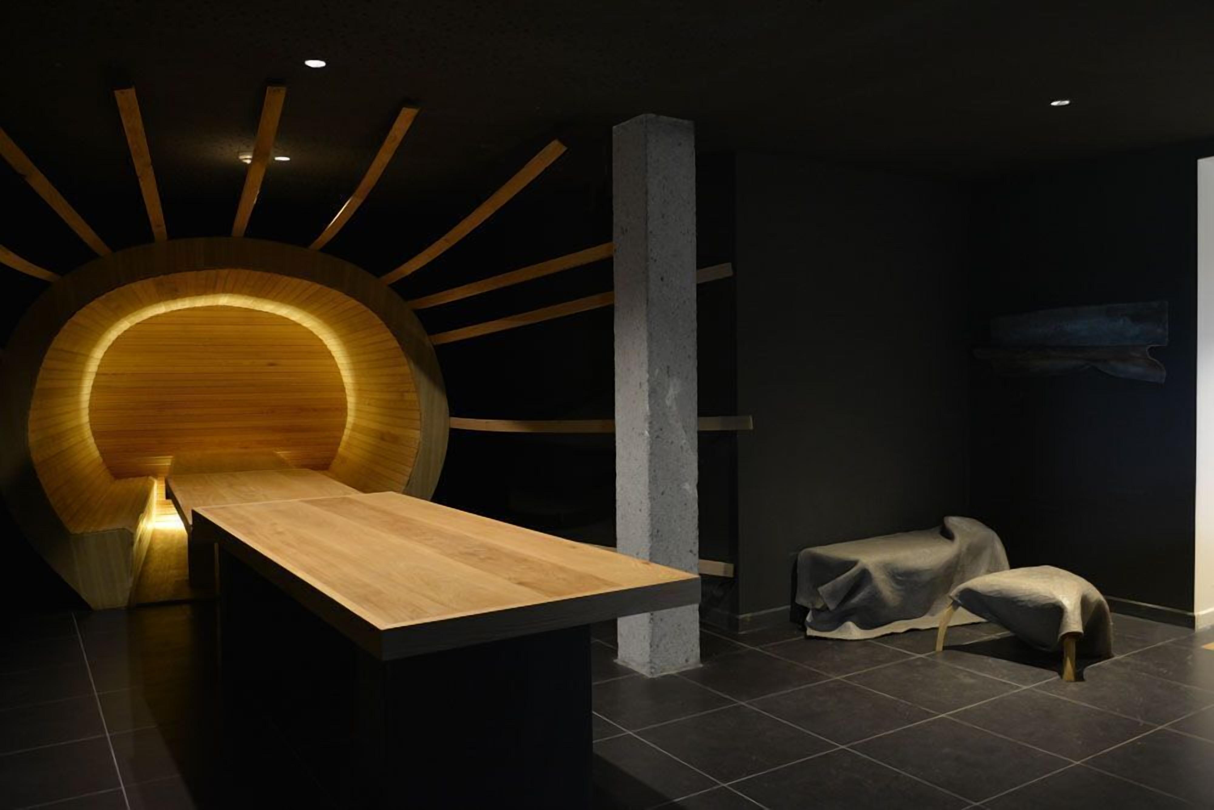 Una habitación moderna con un mostrador de recepción de madera, un elemento decorativo circular de madera en la pared y una pequeña zona de estar con un banco tapizado.