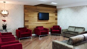 A modern waiting area with red and brown upholstered chairs, a flat-screen TV mounted on a wooden wall, and a potted plant on a glass table. - Hotel Kindermann (Cacador)