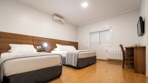 A hotel room with two beds, a wooden headboard, a desk with a chair, a window with blinds, and an air conditioning unit. - Hotel Kindermann (Cacador)